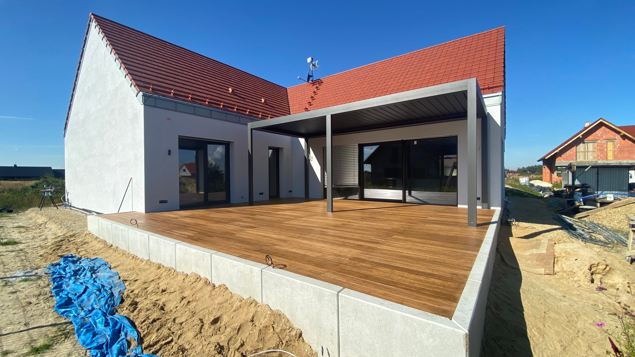 Nowoczesny dom z tarasem z desek kompozytowych i metalową pergolą w tle, widok z poziomu gruntu, błękitne niebo.