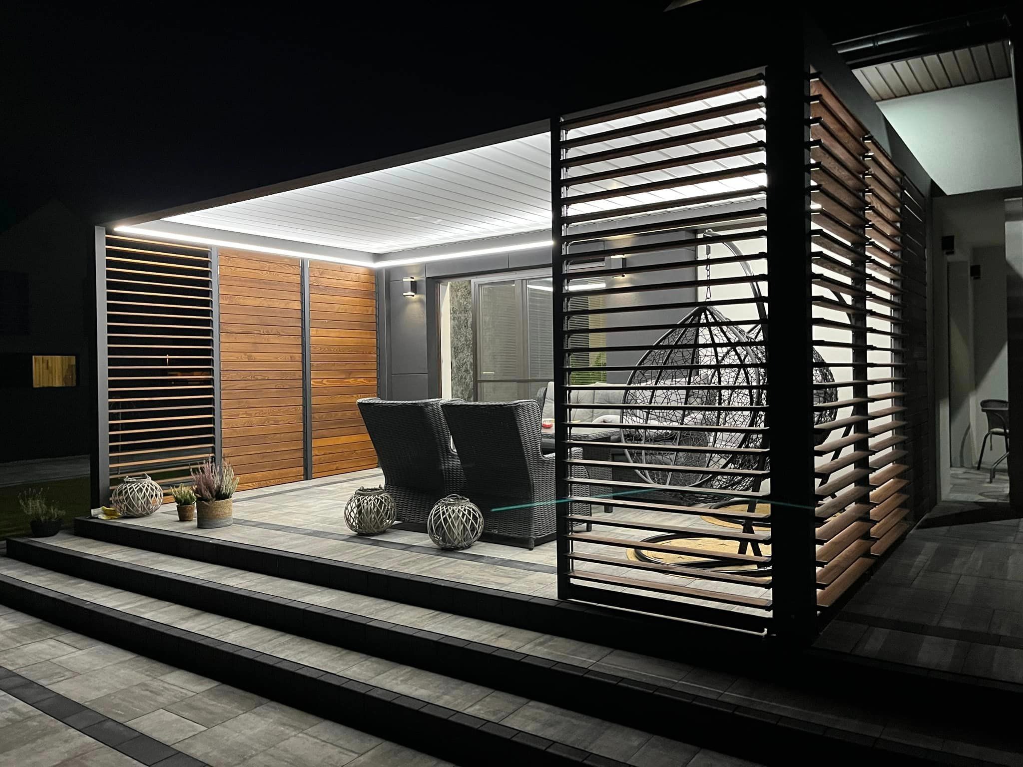 Oświetlona pergola z drewnianymi lamelami i metalową konstrukcją, widoczne meble ogrodowe: dwa fotele i wiszące krzesło, dekoracje roślinne, nocne oświetlenie.