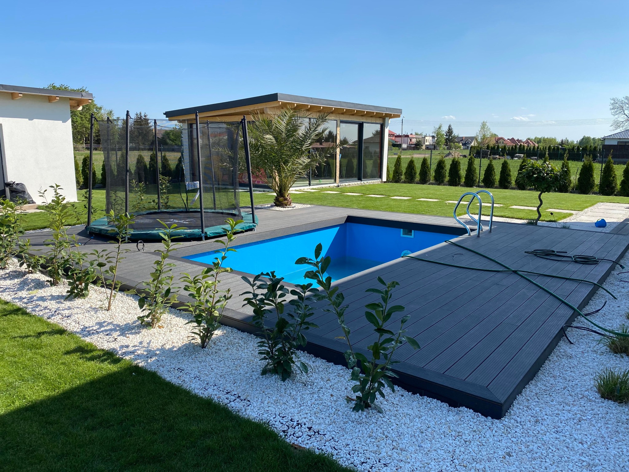 Niebieski basen otoczony szarym tarasem kompozytowym i białymi kamieniami, z widokiem na altanę i trampolinę w zadbanym ogrodzie.