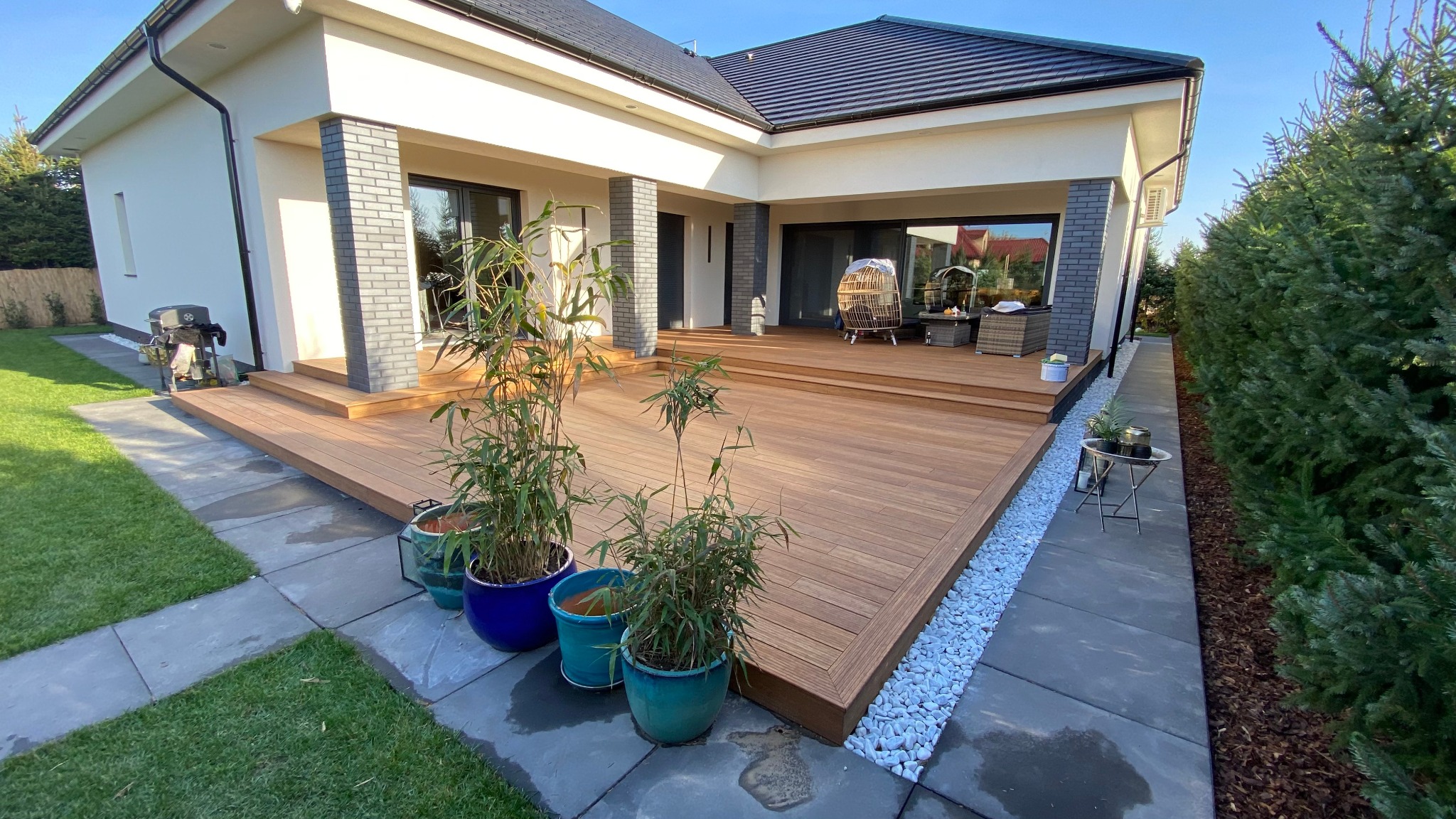 Przestronny taras z desek kompozytowych, z niskimi schodami prowadzącymi do domu, zdobiony donicami z bambusem i białymi kamieniami, z widocznymi meblami ogrodowymi w tle.