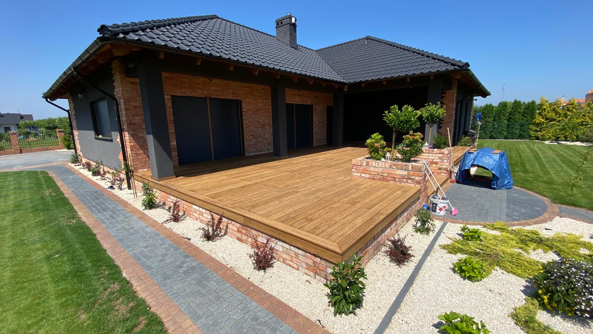 Przestronny taras drewniany z ceglanym obramowaniem, dopasowany do nowoczesnego domu z cegły i ciemną dachówką, w tle zadbany trawnik i błękitne niebo.