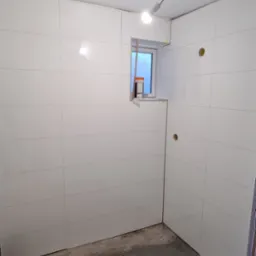 Wnętrze w trakcie wykańczania z białymi, błyszczącymi płytkami na ścianach i suficie, widoczne otwory na instalacje elektryczne i okno z pomarańczowym pojemnikiem na parapecie, surowa podłoga...