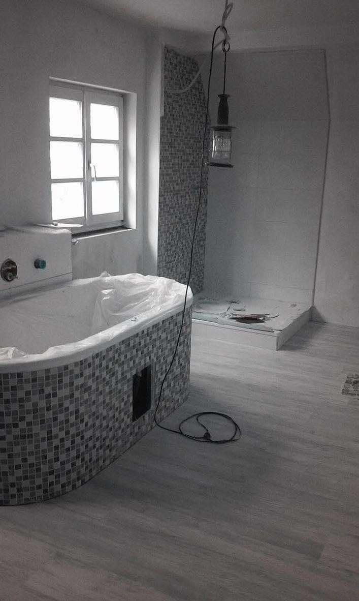 Niedokończona łazienka z wanną w mozaice zabezpieczoną folią, kabiną prysznicową w budowie i lampą warsztatową zwisającą z sufitu.