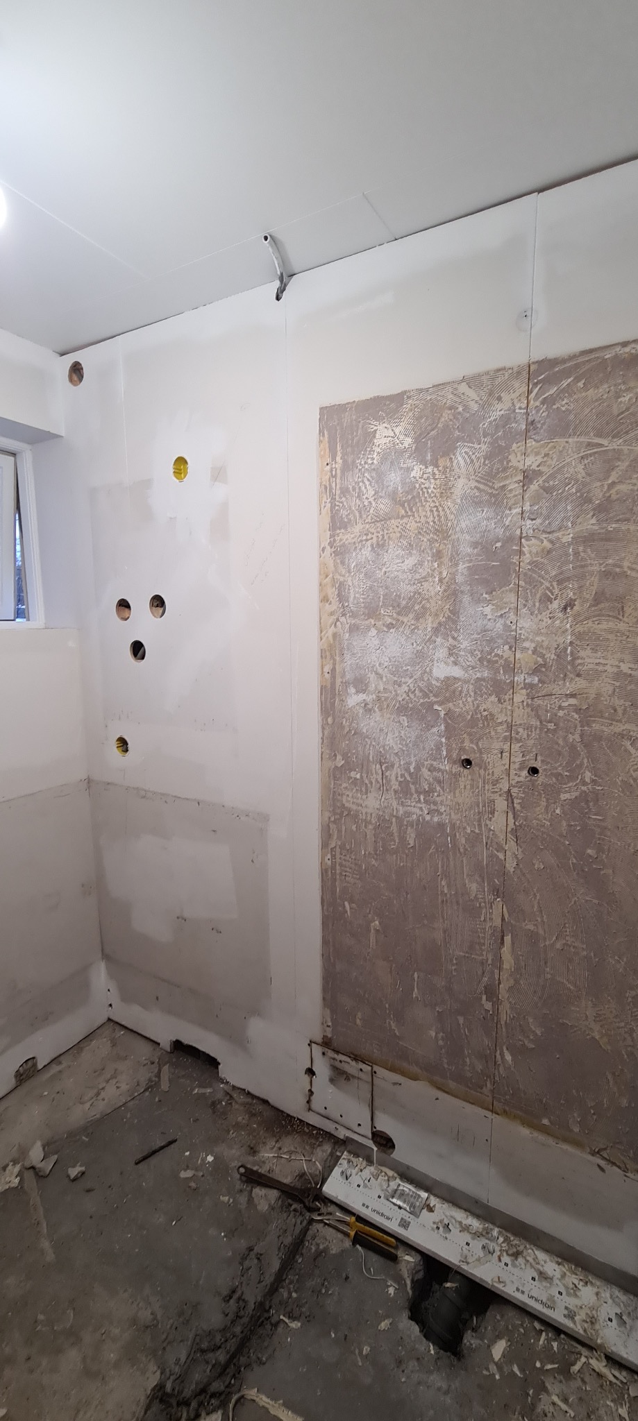 Wnętrze w trakcie prac przygotowawczych: odkryte ściany z widocznymi śladami po starej okładzinie, wywiercone otwory na instalacje, surowy beton na podłodze, narzędzia i listwa przypodłogowa leżące...