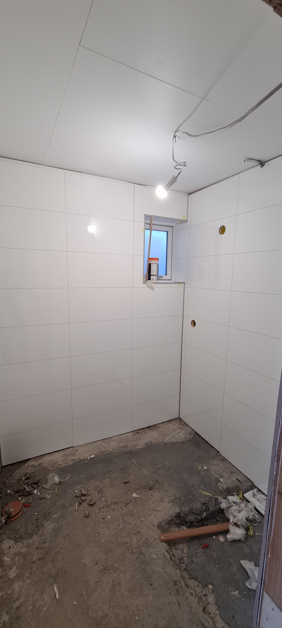 Wnętrze w trakcie wykańczania z białymi, błyszczącymi płytkami na ścianach i suficie, widoczne otwory na instalacje elektryczne i okno z pomarańczowym pojemnikiem na parapecie, surowa podłoga...