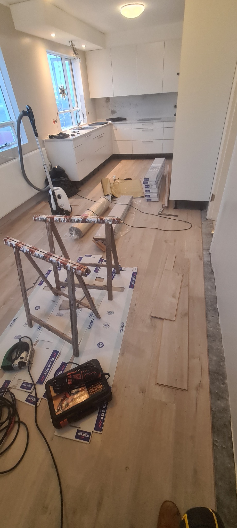 Wnętrze kuchni w trakcie układania jasnych paneli podłogowych, widoczne stojaki malarskie, paczki paneli, odkurzacz, skrzynka z narzędziami oraz niedokończona podłoga.