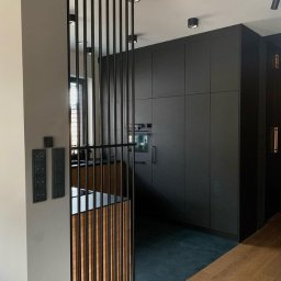 MEBLEWOOD - Nowoczesna kuchnia z zabudową w ciemnych odcieniach, oddzielona ażurową ścianką działową z pionowych listew z elementami drewna, widoczne włączniki na ścianie.