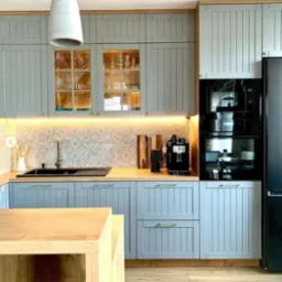 Jasnoszara kuchnia z drewnianym blatem, frezowanymi frontami, oświetleniem LED pod szafkami wiszącymi i wyspą kuchenną z blatem z litego drewna.