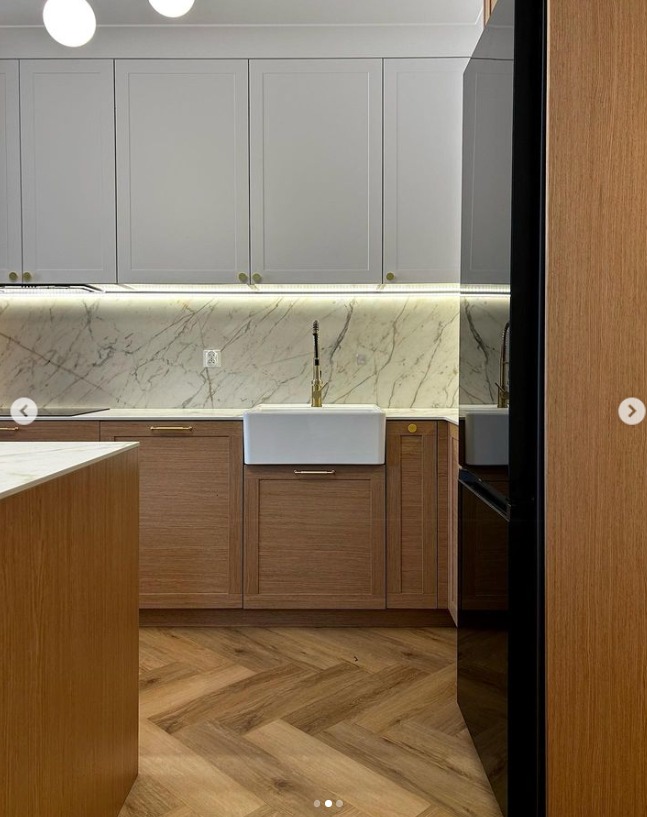 Nowoczesna kuchnia z białymi górnymi szafkami, drewnianymi dolnymi szafkami i marmurowym backsplashiem, akcentowana złotą armaturą i oświetleniem LED pod szafkami, z drewnianą podłogą ułożoną...