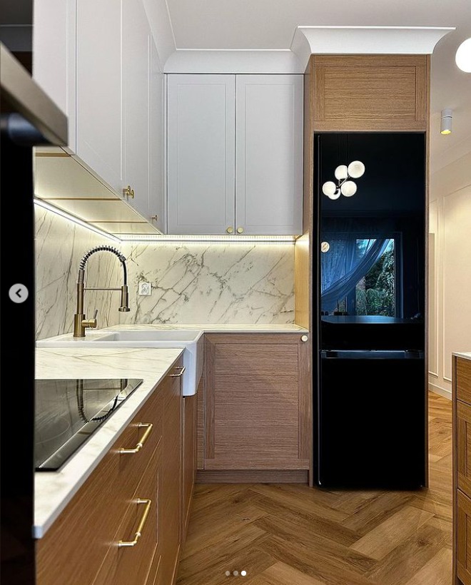 Nowoczesna kuchnia z drewnianymi frontami szafek dolnych i białymi górnymi, marmurowym backsplashiem, złotą armaturą i czarną lodówką, parkiet ułożony w jodełkę.