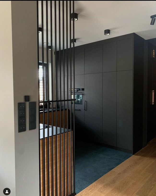 Nowoczesna kuchnia z zabudową w ciemnych odcieniach, oddzielona ażurową ścianką działową z pionowych listew z elementami drewna, widoczne włączniki na ścianie.