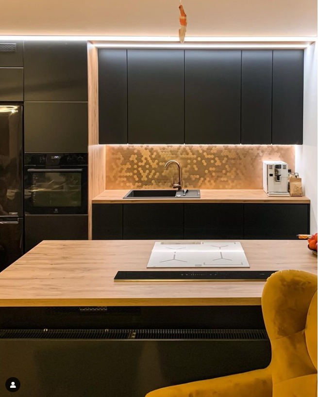 Nowoczesna, czarna kuchnia z drewnianym blatem i miedzianą mozaiką nad zlewem. Na wyspie kuchennej wbudowana płyta indukcyjna. W tle widoczny fragment żółtego fotela.