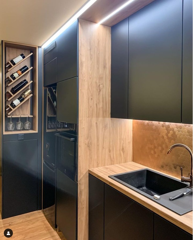 Nowoczesna kuchnia z czarnymi frontami, drewnopodobnymi wstawkami i podświetleniem LED, z wbudowaną szafką na wino i złotą mozaiką nad blatem.