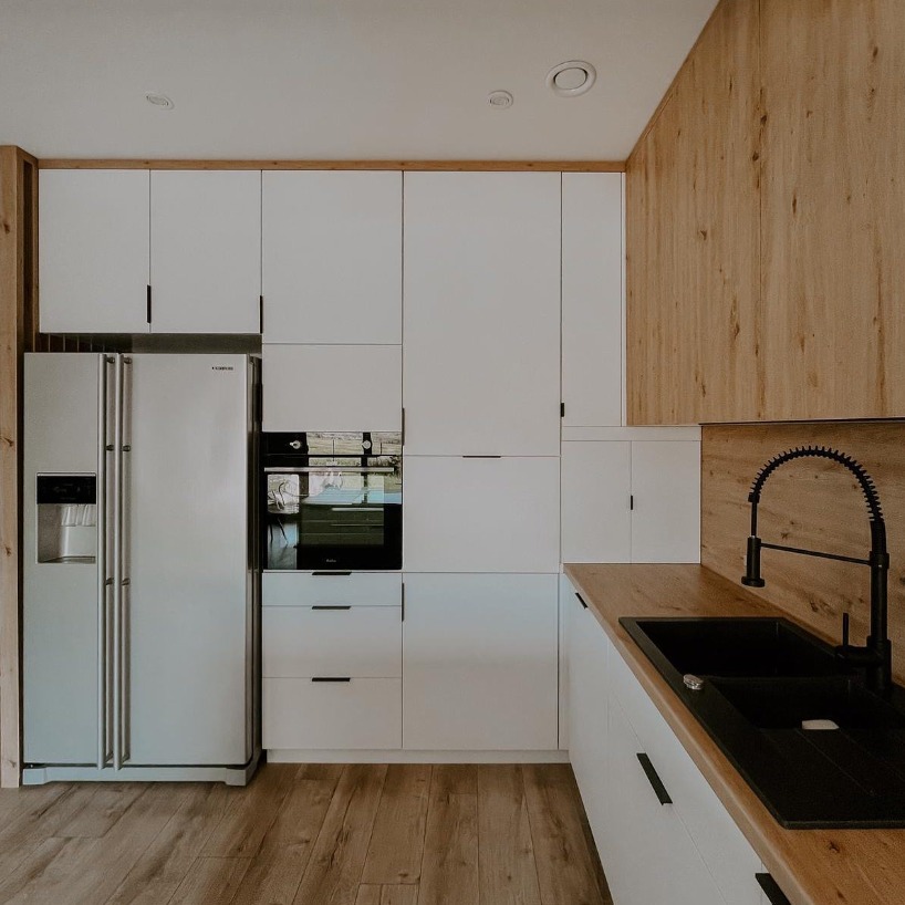 Nowoczesna, minimalistyczna kuchnia z białymi frontami szafek, drewnianym blatem i elementami wykończenia, czarną armaturą i dużym, dwudrzwiowym, białym lodówką.
