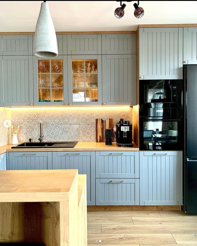 Jasnoszara kuchnia z drewnianym blatem, frezowanymi frontami, oświetleniem LED pod szafkami wiszącymi i wyspą kuchenną z blatem z litego drewna.