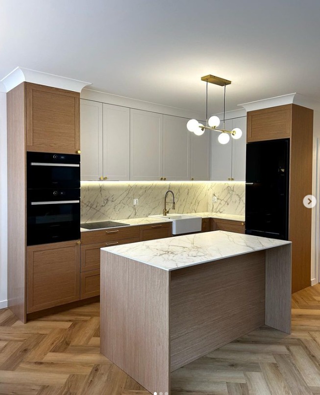 Nowoczesna kuchnia z wyspą, fronty szafek w odcieniach bieli i drewna, marmurowy blat i backsplash, oświetlenie punktowe pod szafkami wiszącymi, parkiet w jodełkę na podłodze.