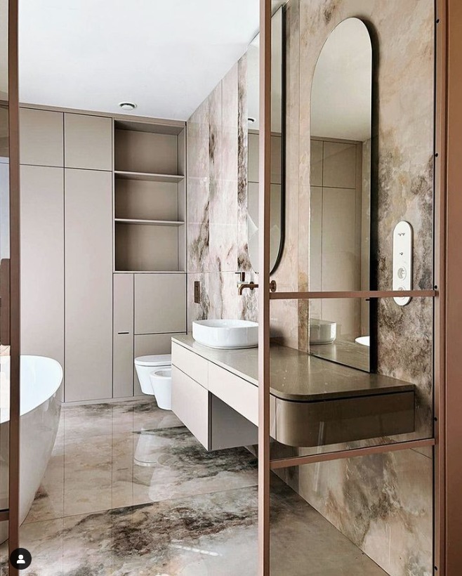 Elegancka łazienka w odcieniach beżu z marmurkową płytką, owalnym lustrem i minimalistyczną armaturą, widok zza przeszklonej ścianki.