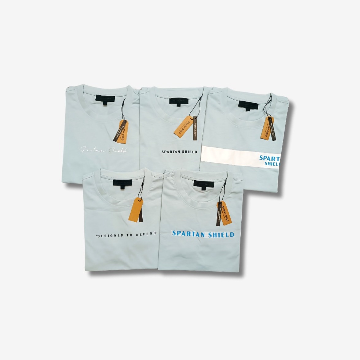 Jasnoniebieskie koszulki z nadrukami 'Spartan Shield' i zawieszkami, ułożone na białym tle. Różne warianty graficzne nadruków.