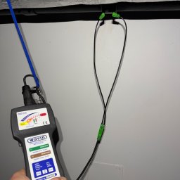 LuLu Instal Bohdan Pasevych - Pomiar zgrzewu rur elektrooporowych przy użyciu zgrzewarki Wavin Duo 315. Widoczne przewody i urządzenie pomiarowe na tle szarej ściany i izolowanej rury.