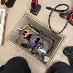 LuLu Instal Bohdan Pasevych - Otwarty właz rewizyjny z zanurzoną pompą, widoczne narzędzia i fragment osoby w czarnych spodniach. Prace hydrauliczne w trakcie realizacji, z bliska.