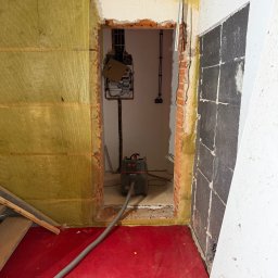 LuLu Instal Bohdan Pasevych - Wnętrze w trakcie remontu: ściany z izolacją, odsłonięta cegła, instalacja elektryczna i piec. Na podłodze czerwona wykładzina i odkurzacz budowlany Starmix.