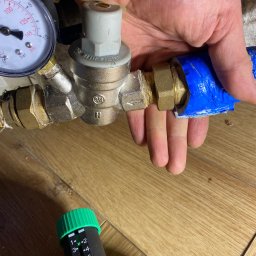 LuLu Instal Bohdan Pasevych - Instalacja wodna: zawór redukcyjny ciśnienia z manometrem, podłączony do rury owiniętej niebieską taśmą. Widoczne złącza mosiężne i regulator z zielonym pokrętłem.