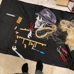 LuLu Instal Bohdan Pasevych - Mosiężne złączki hydrauliczne, klucze i narzędzia na czarnej macie z motywem czaszki. Widoczne stopy w skarpetkach i spodniach. Przygotowanie do montażu instalacji.