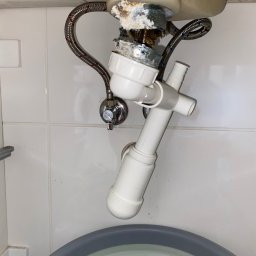 LuLu Instal Bohdan Pasevych - Nieszczelny syfon umywalki z widocznymi śladami korozji i wycieków, pod spodem wiadro. Ujęcie z dołu, na białym tle płytek.