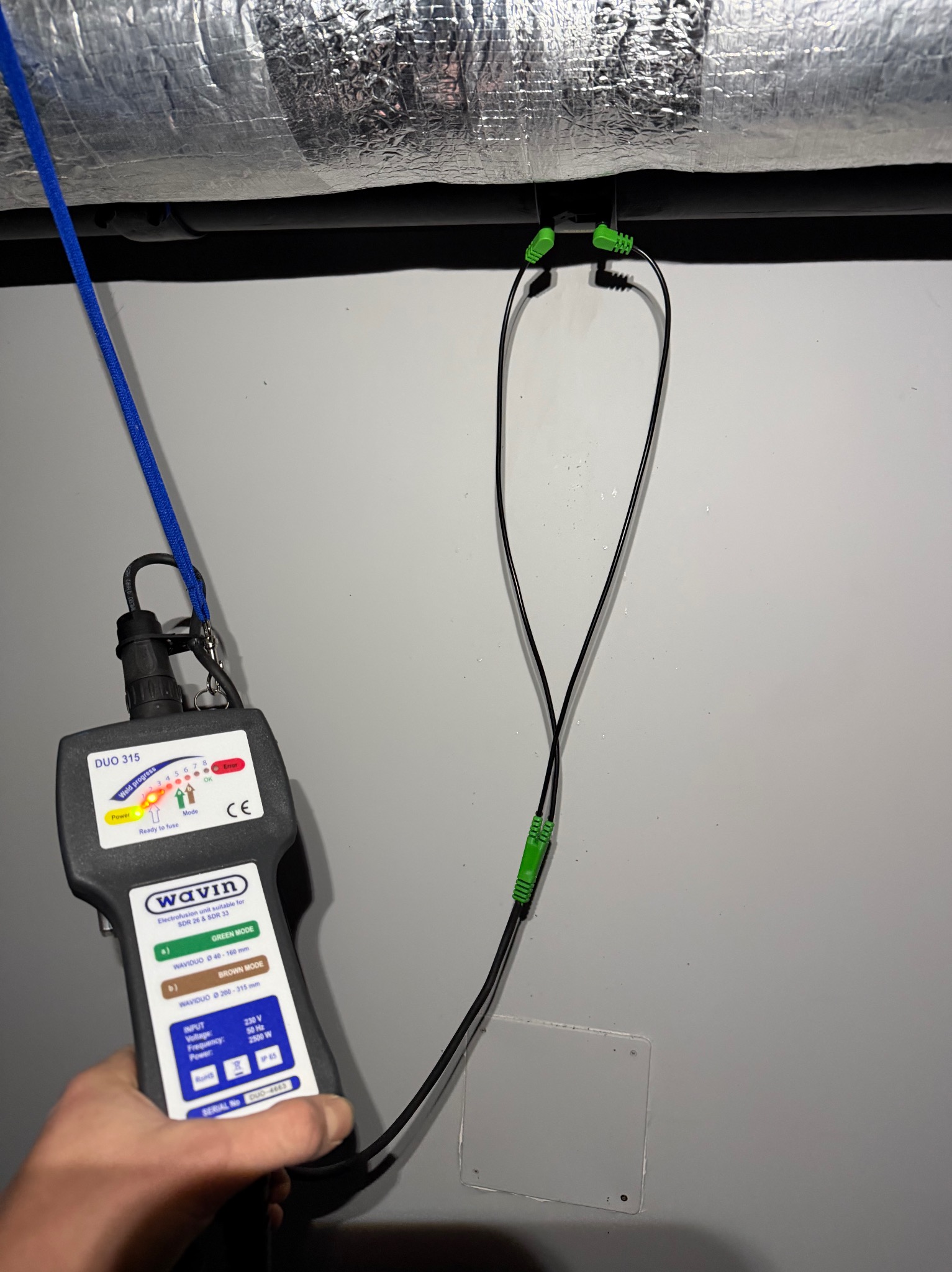Pomiar zgrzewu rur elektrooporowych przy użyciu zgrzewarki Wavin Duo 315. Widoczne przewody i urządzenie pomiarowe na tle szarej ściany i izolowanej rury.