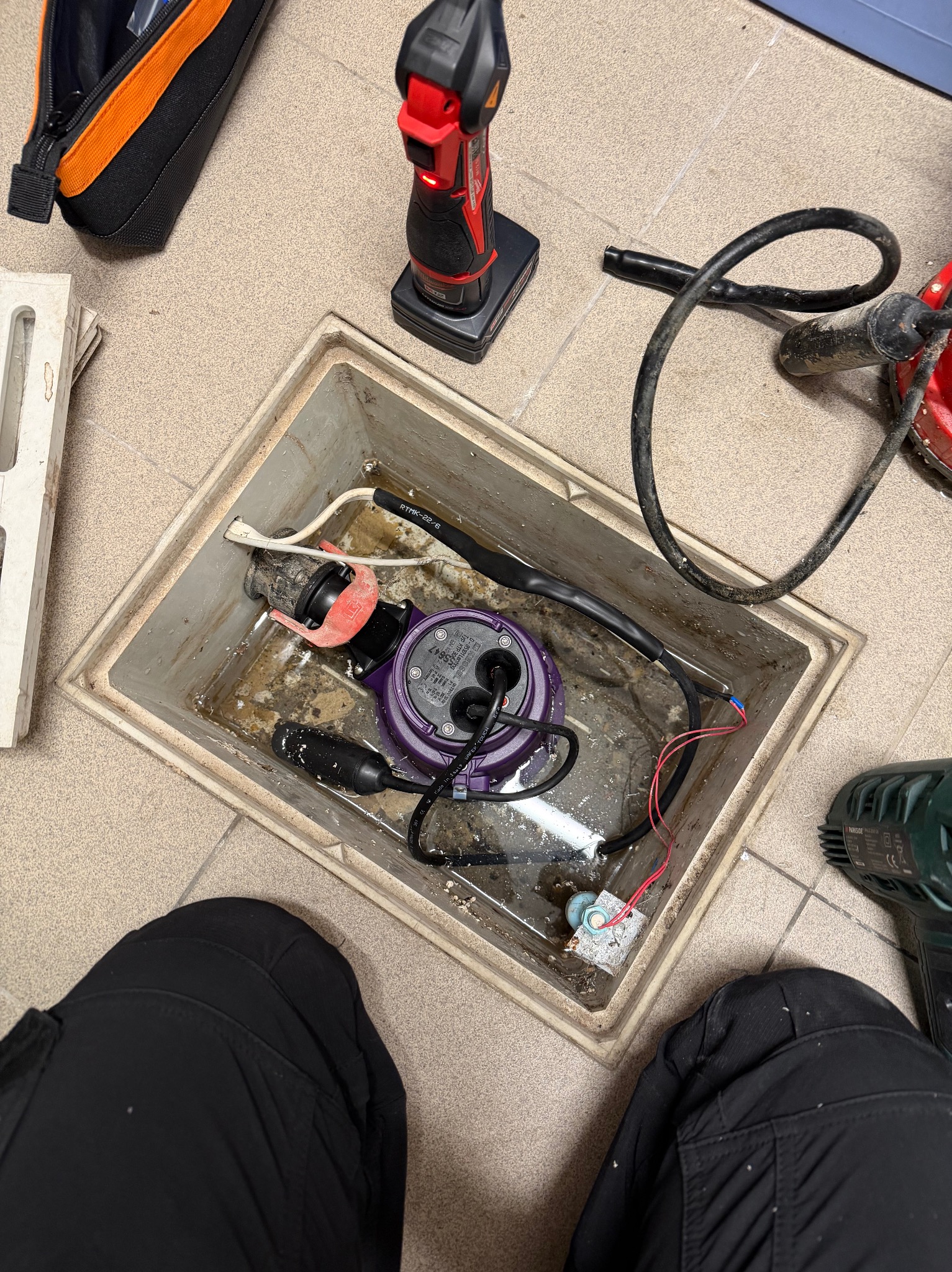 Otwarty właz rewizyjny z zanurzoną pompą, widoczne narzędzia i fragment osoby w czarnych spodniach. Prace hydrauliczne w trakcie realizacji, z bliska.