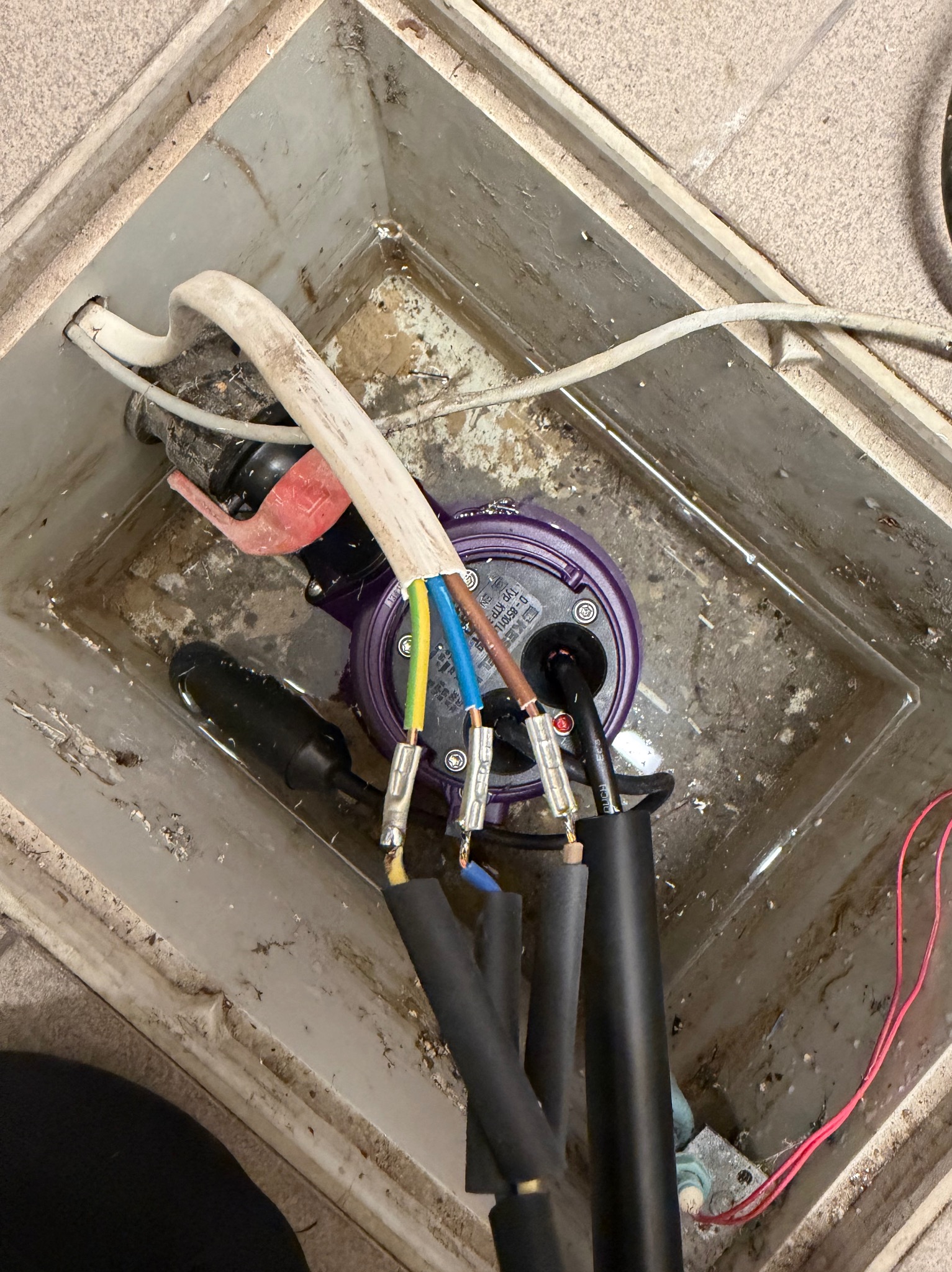 Podłączenie kabli do pompy zanurzeniowej w studzience. Widoczne przewody z kolorowymi izolacjami i czarne rury. Praca instalacyjna w szarej, betonowej studzience.