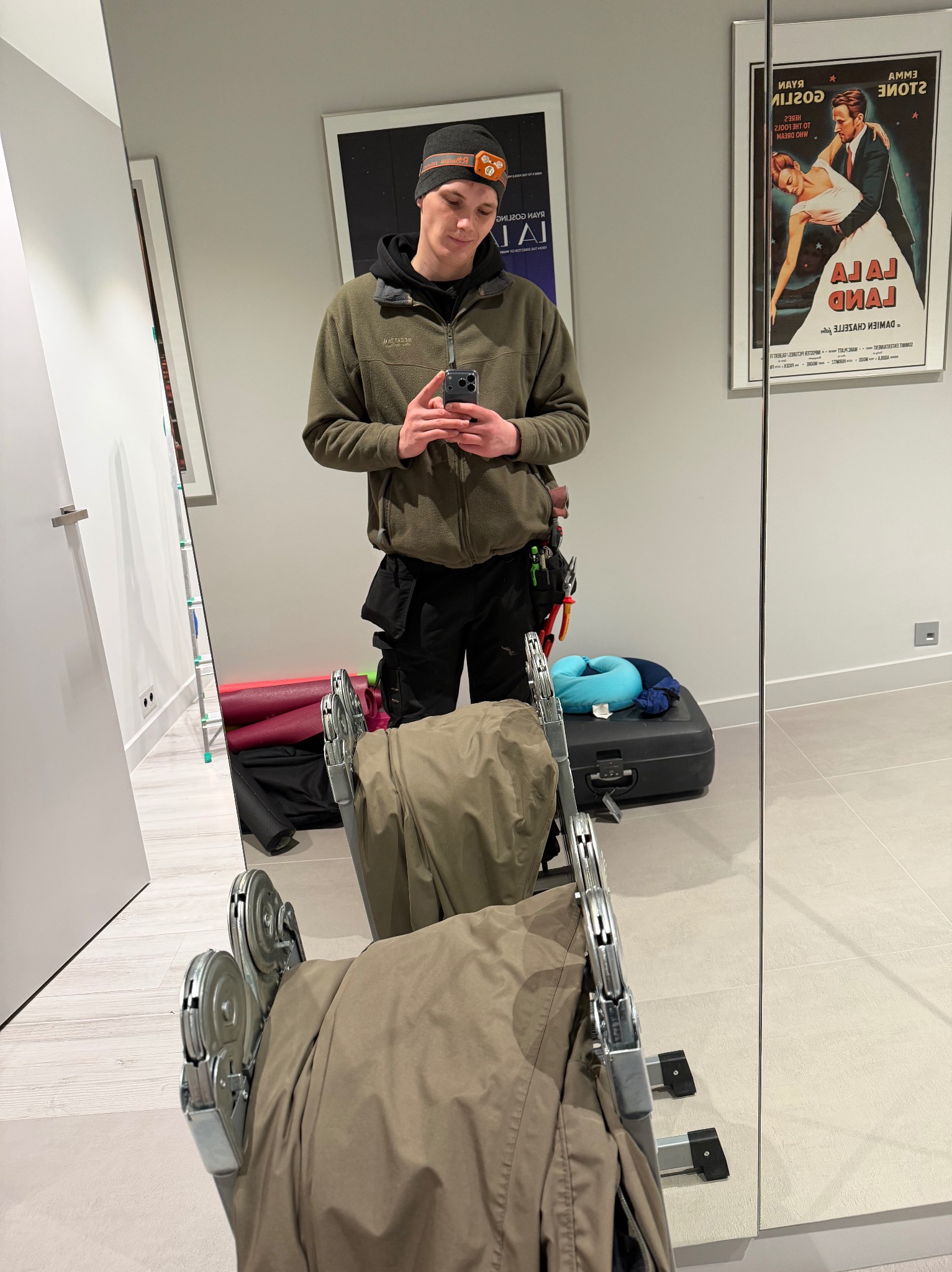 Elektryk w odzieży roboczej robi selfie w lustrze, widoczne narzędzia i drabina. W tle plakat filmowy 'La La Land' i inne elementy wyposażenia wnętrza.