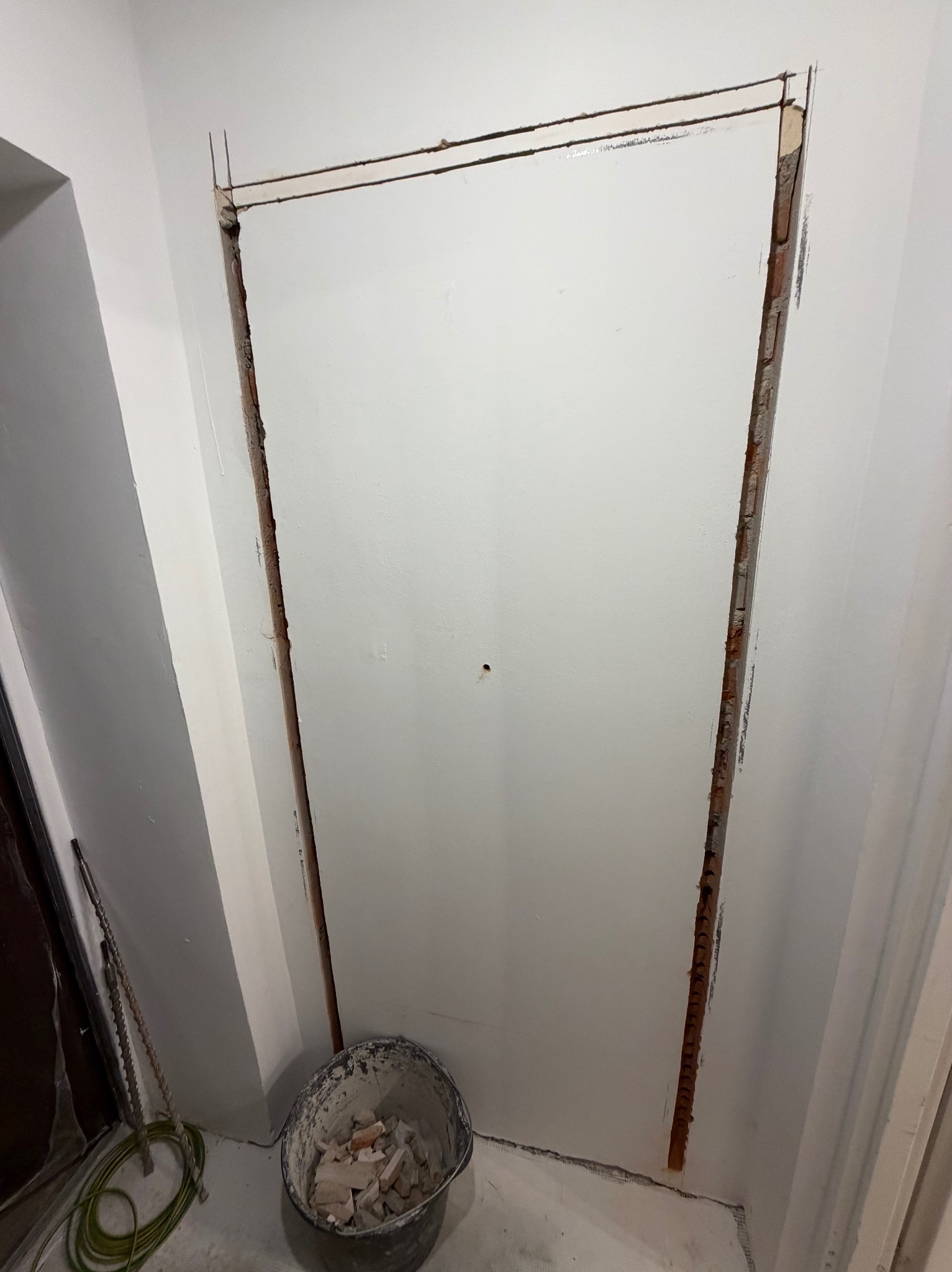 Przygotowany otwór w ścianie pod montaż ościeżnicy, widoczne ślady po kuciu i resztki gruzu w wiadrze. Prace remontowe w trakcie realizacji.