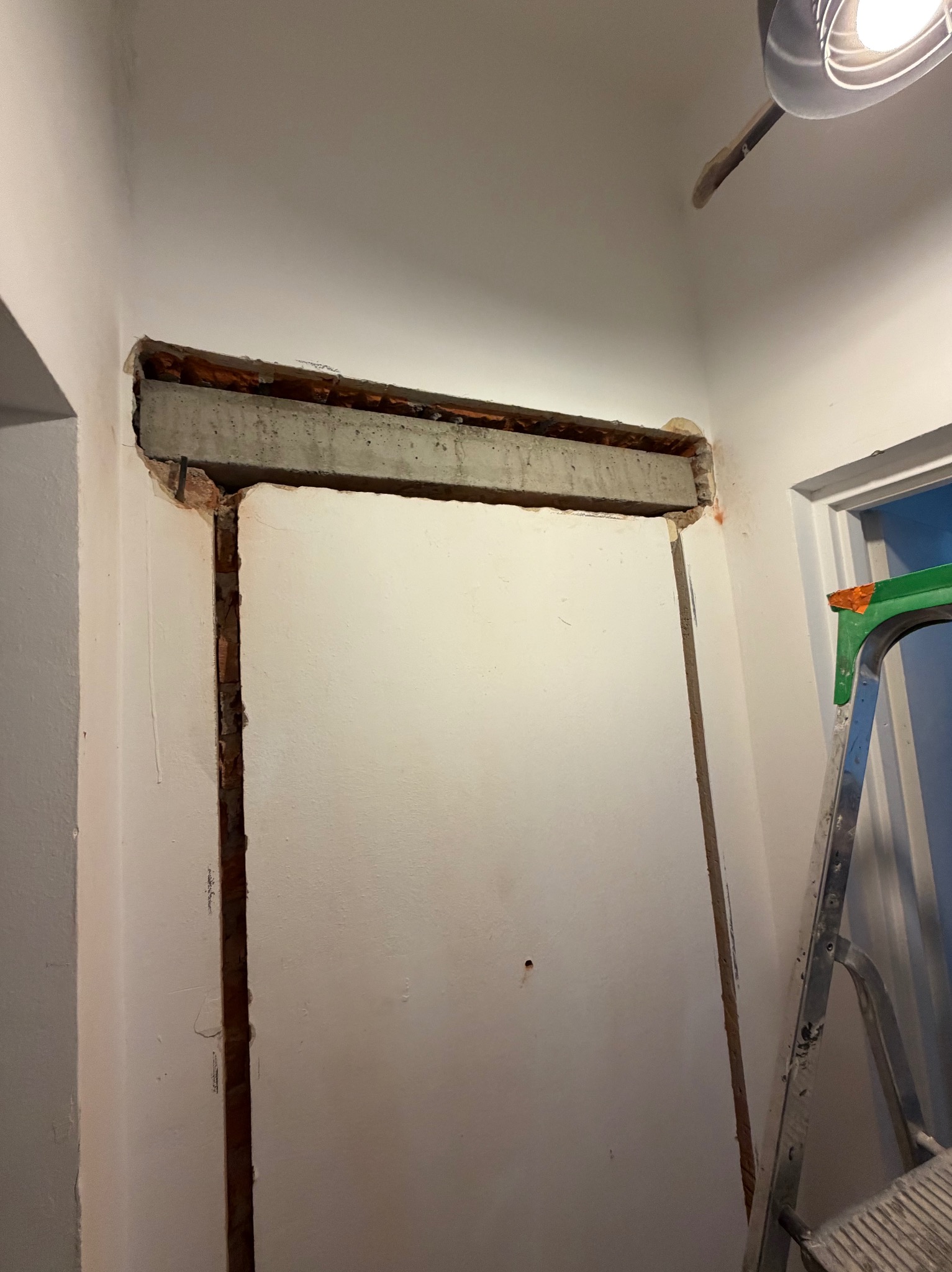 Otwór drzwiowy w trakcie prac remontowych, widoczny betonowy nadproże i fragment ściany z cegły. W tle fragment drabiny malarskiej i lampy sufitowej.