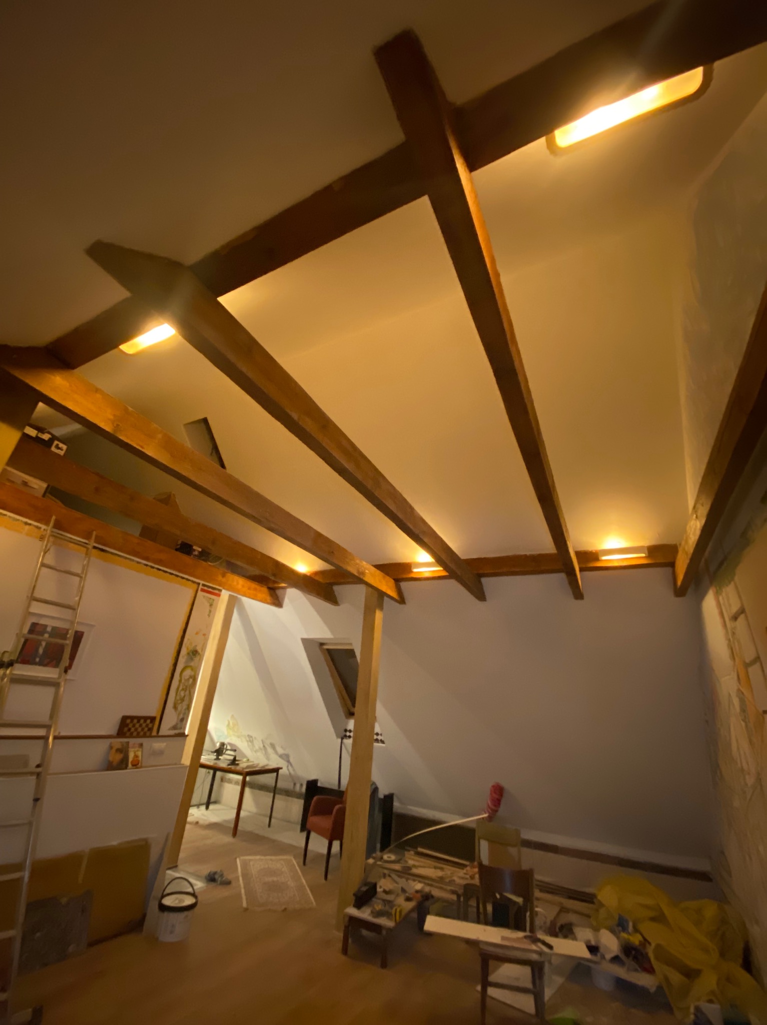 Strych z drewnianymi belkami stropowymi i zamontowanym oświetleniem. Widoczna drabina, narzędzia i materiały budowlane. Ujęcie z dołu, szeroki kąt.