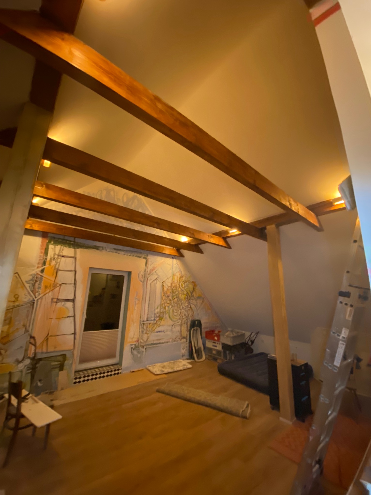 Podświetlane drewniane belki stropowe w pokoju na poddaszu z malowidłami na ścianach. Widoczny fragment drabiny i wyposażenia.