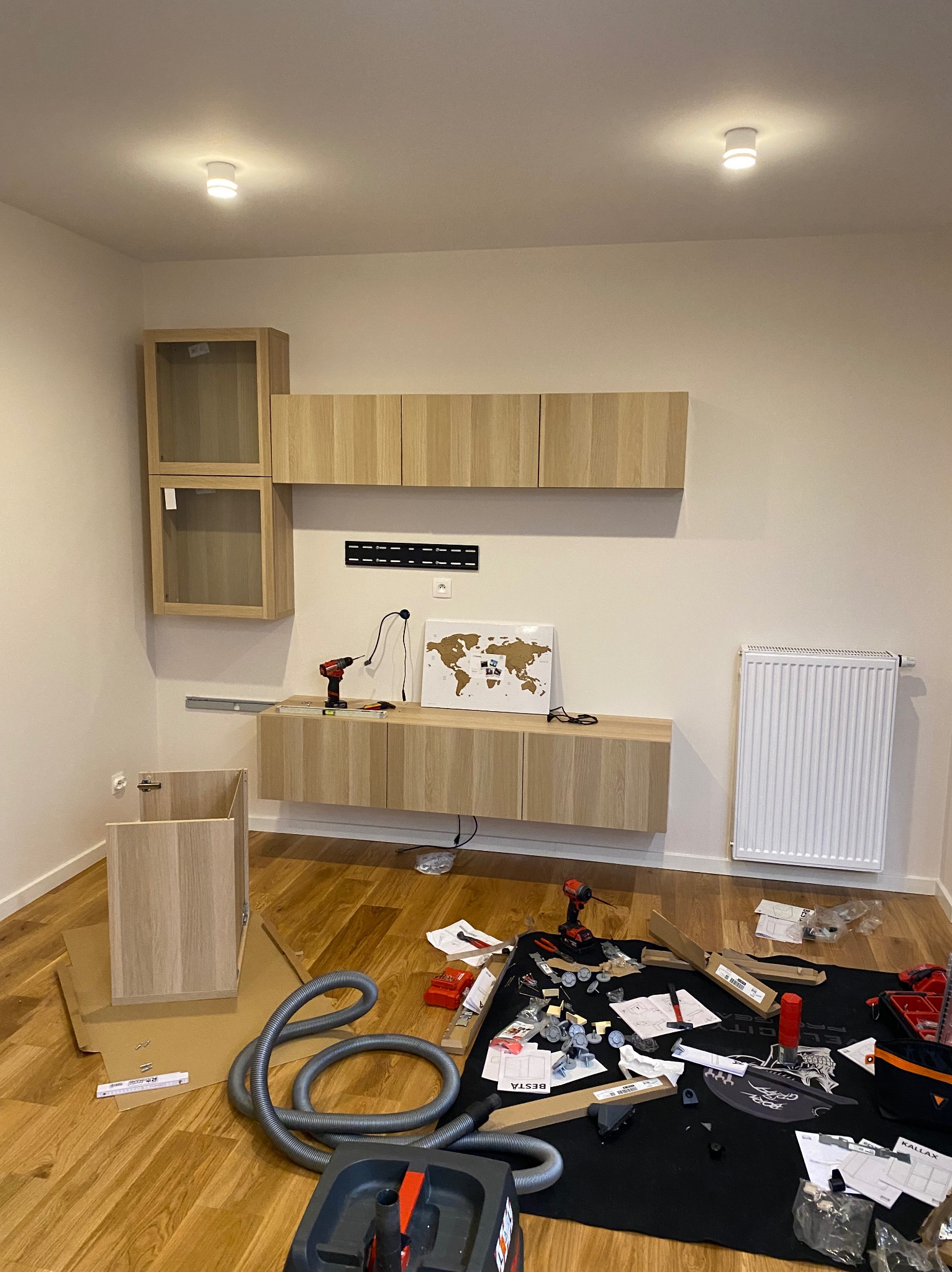 Montaż mebli modułowych w salonie: wiszące szafki z jasnego drewna, narzędzia, odkurzacz i elementy montażowe na podłodze. Widoczny grzejnik i mapa świata na ścianie.