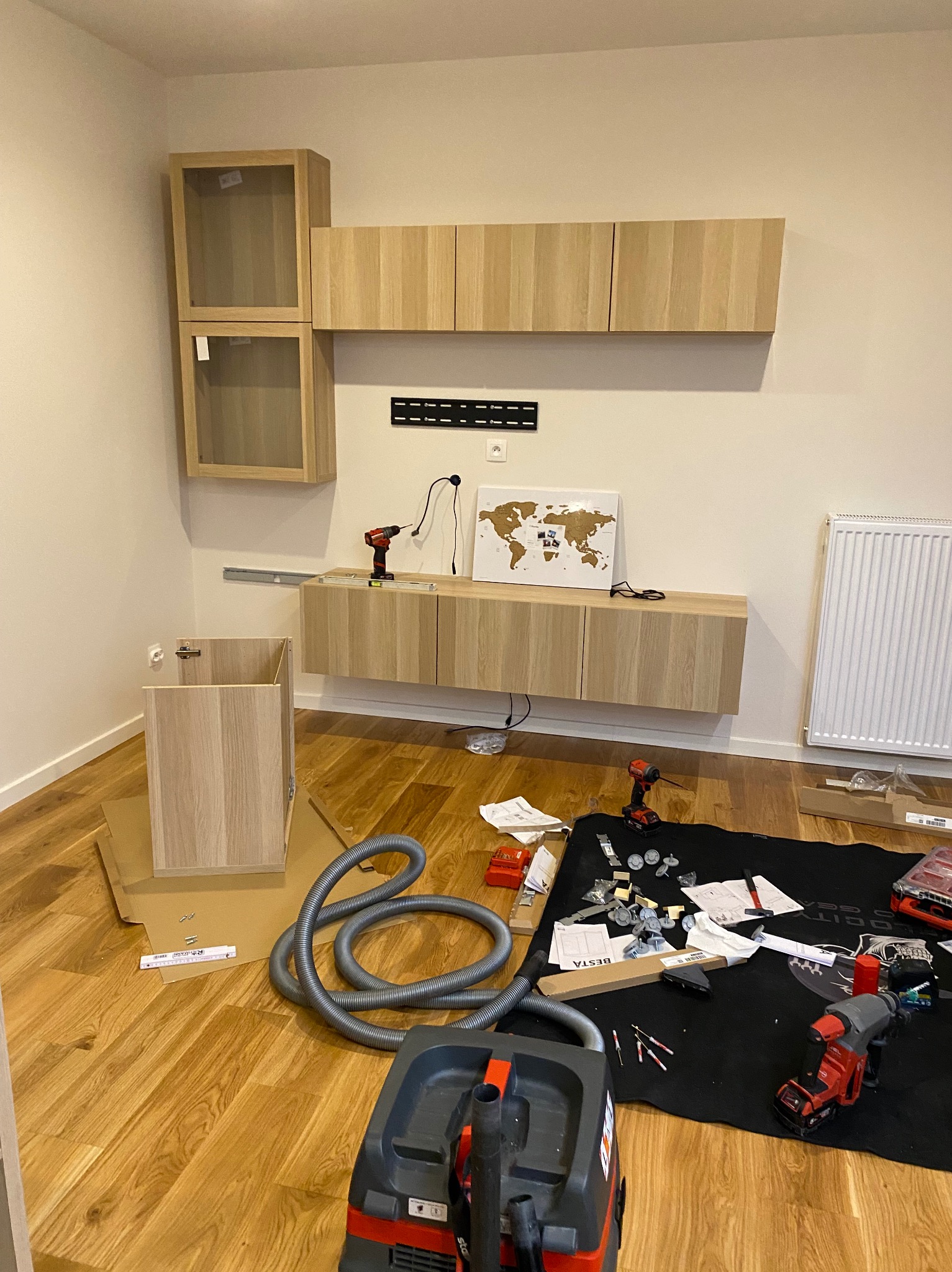 Montaż mebli BESTA: wiszące szafki z jasnego drewna na ścianie, narzędzia i odkurzacz na podłodze. Etap składania, widoczne elementy montażowe i instrukcja.
