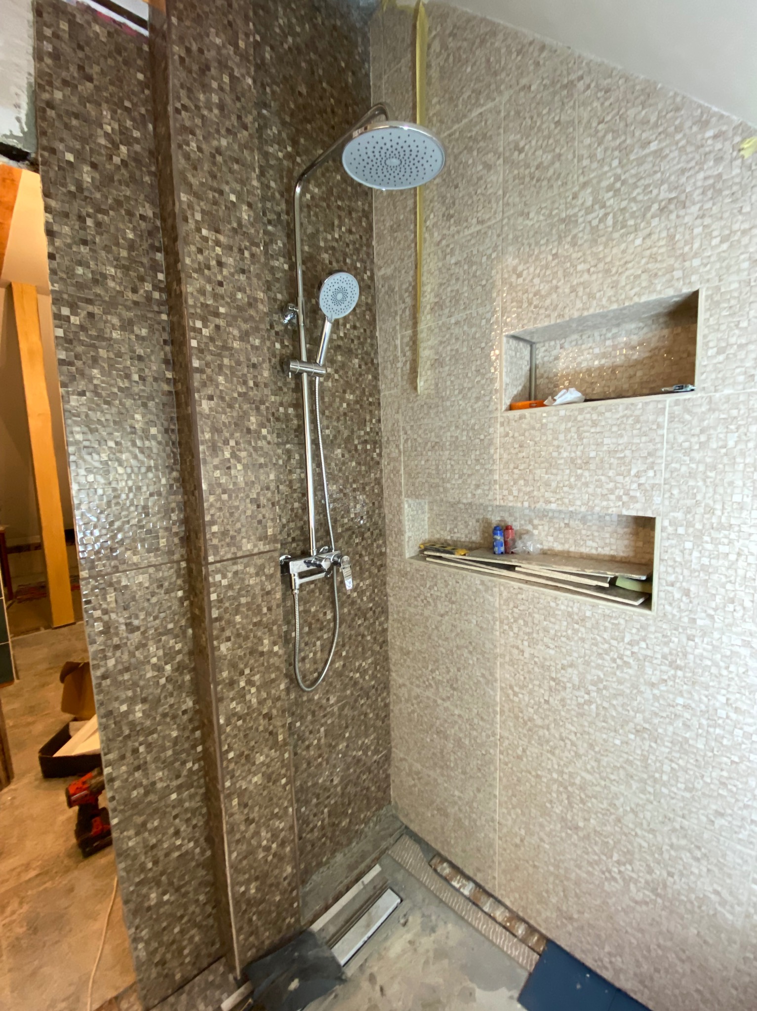 Wykończona kabina prysznicowa z mozaiką w odcieniach brązu i beżu, z wnękami na kosmetyki. Widoczna bateria i deszczownica. Częściowo widoczne narzędzia.