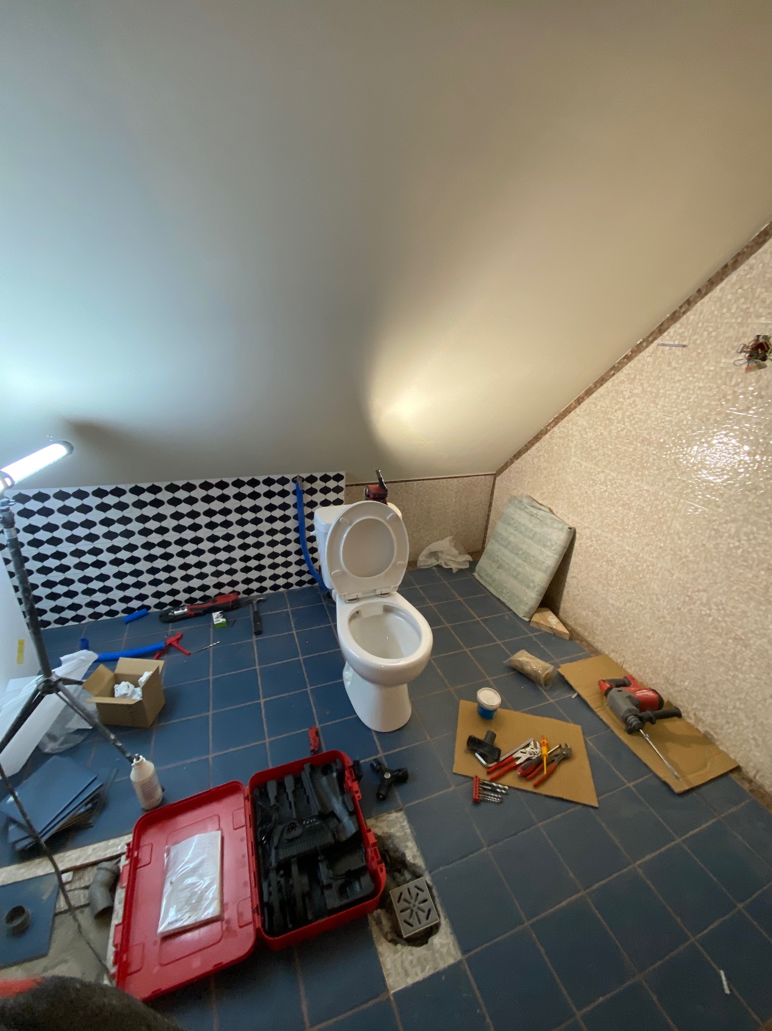 Montaż toalety w łazience na poddaszu z niebieską podłogą w kratkę. Widoczne narzędzia i otwarty sedes. Ukończony etap prac hydraulicznych.