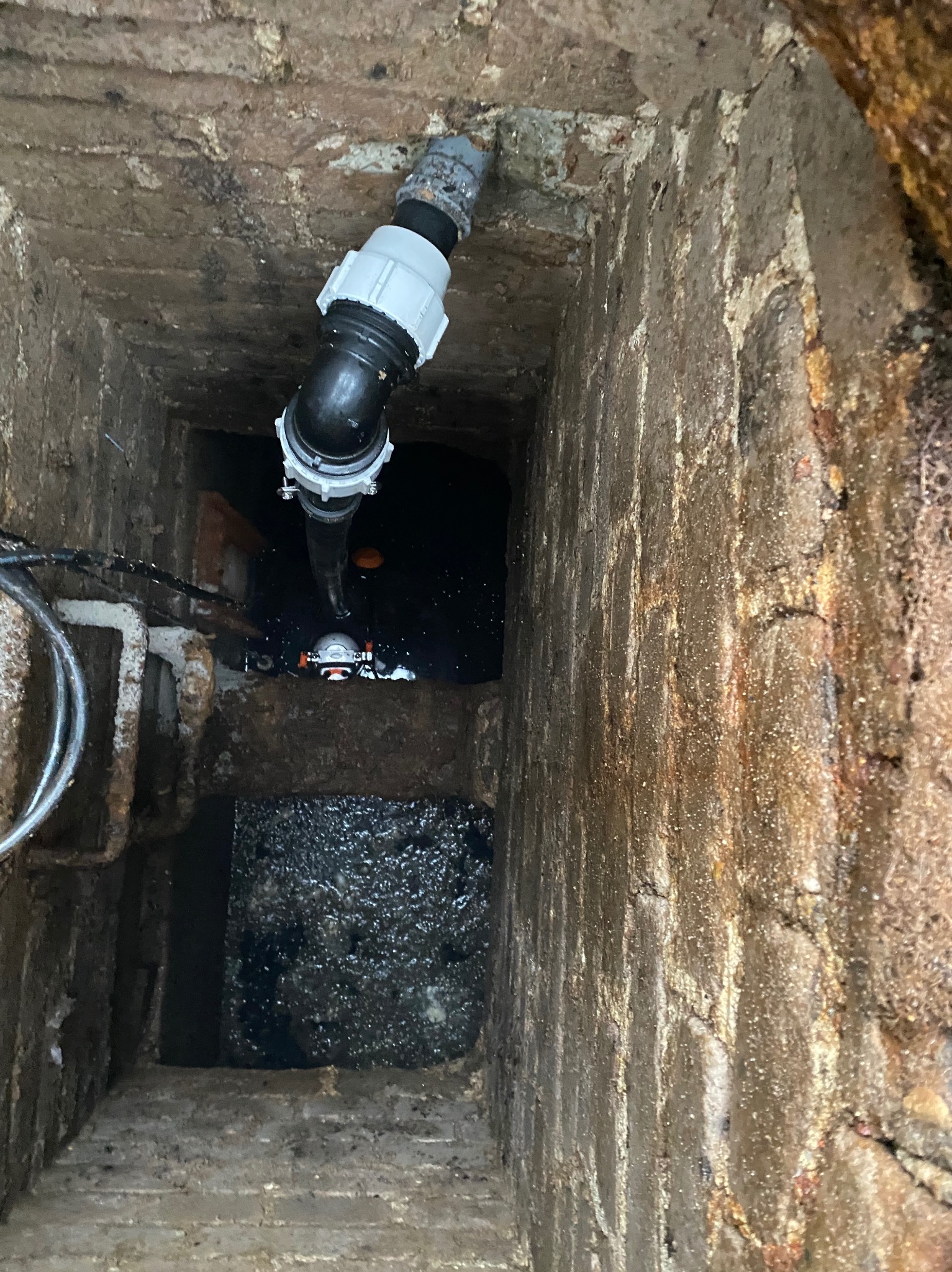 Wnętrze studzienki kanalizacyjnej z rurami PCV i wodą. Widoczne ceglane ściany i dno studzienki. Ujęcie z góry, ukazujące instalację w trakcie prac lub po naprawie.