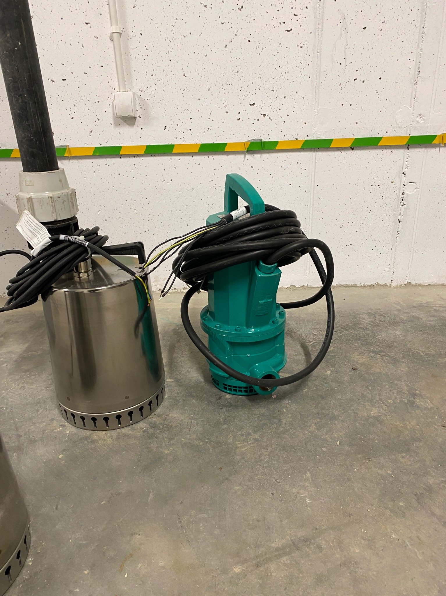 Dwie pompy zanurzeniowe: stalowa i turkusowa, z kablami, na szarym betonie. Gdańsk. Widoczne elementy instalacji elektrycznej na ścianie.