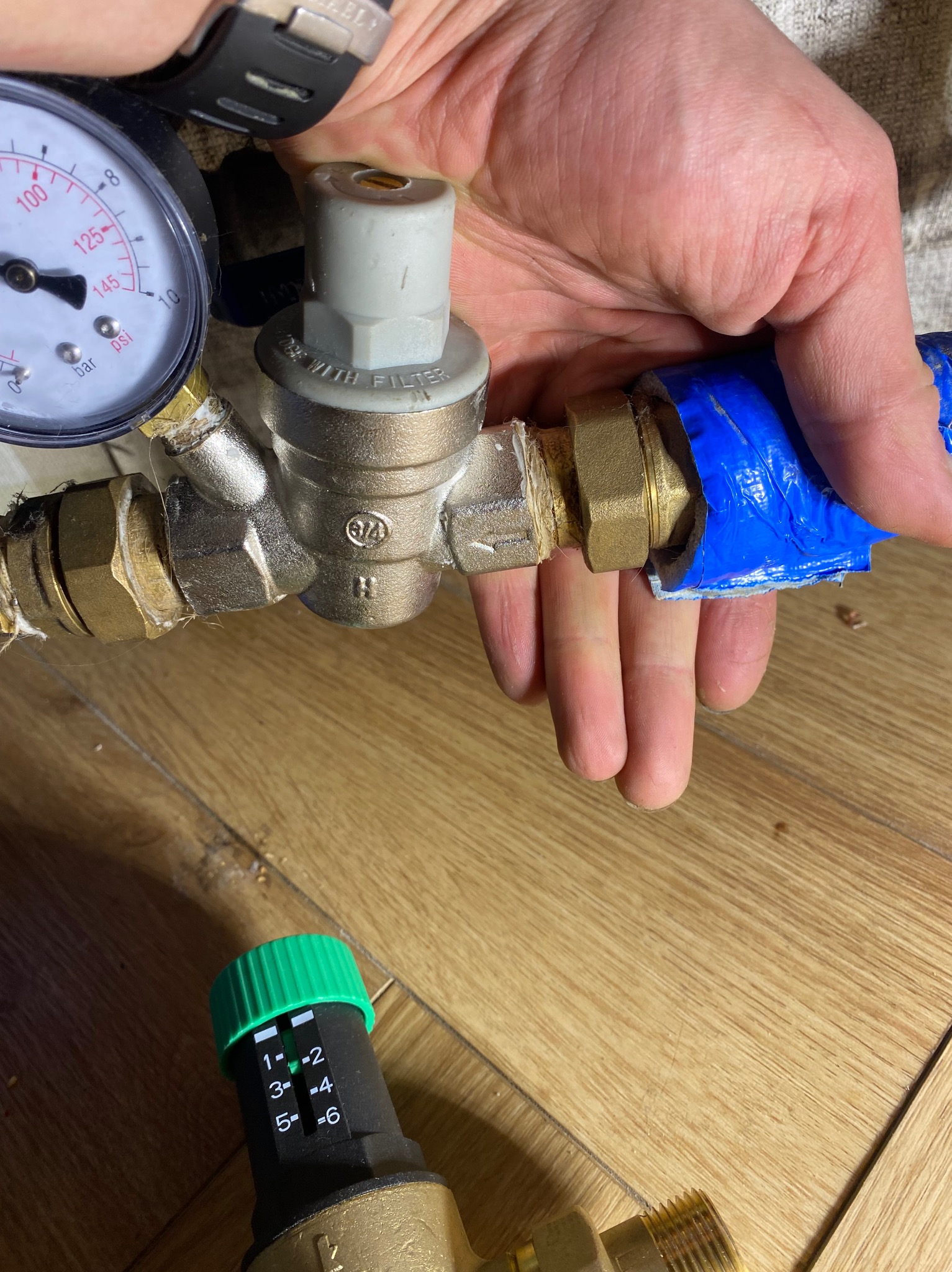 Instalacja wodna: zawór redukcyjny ciśnienia z manometrem, podłączony do rury owiniętej niebieską taśmą. Widoczne złącza mosiężne i regulator z zielonym pokrętłem.