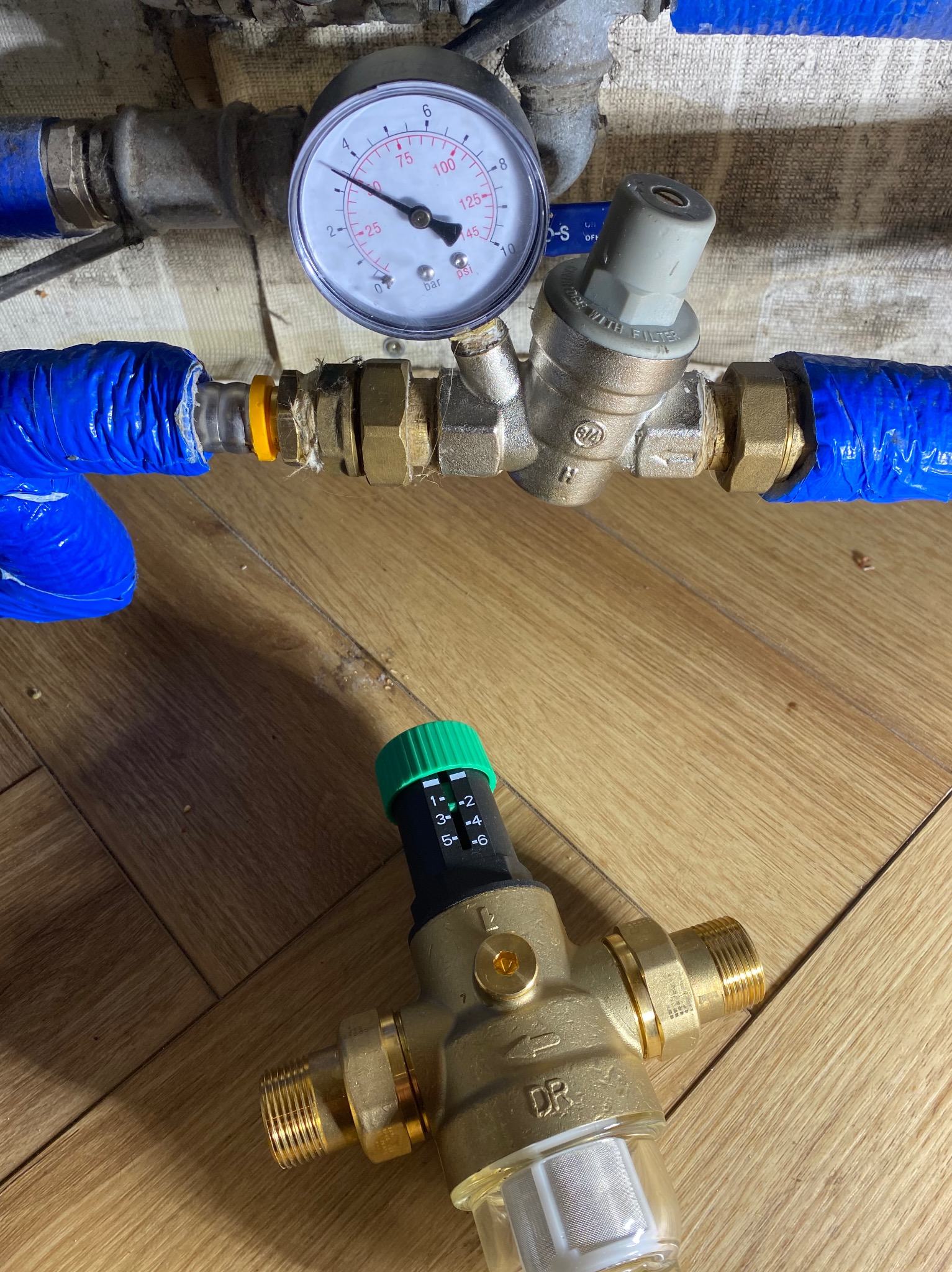 Zawór redukcyjny ciśnienia z manometrem na tle drewnianej podłogi. Element instalacji hydraulicznej z niebieską izolacją na rurach. Widoczny filtr i skala regulacji.