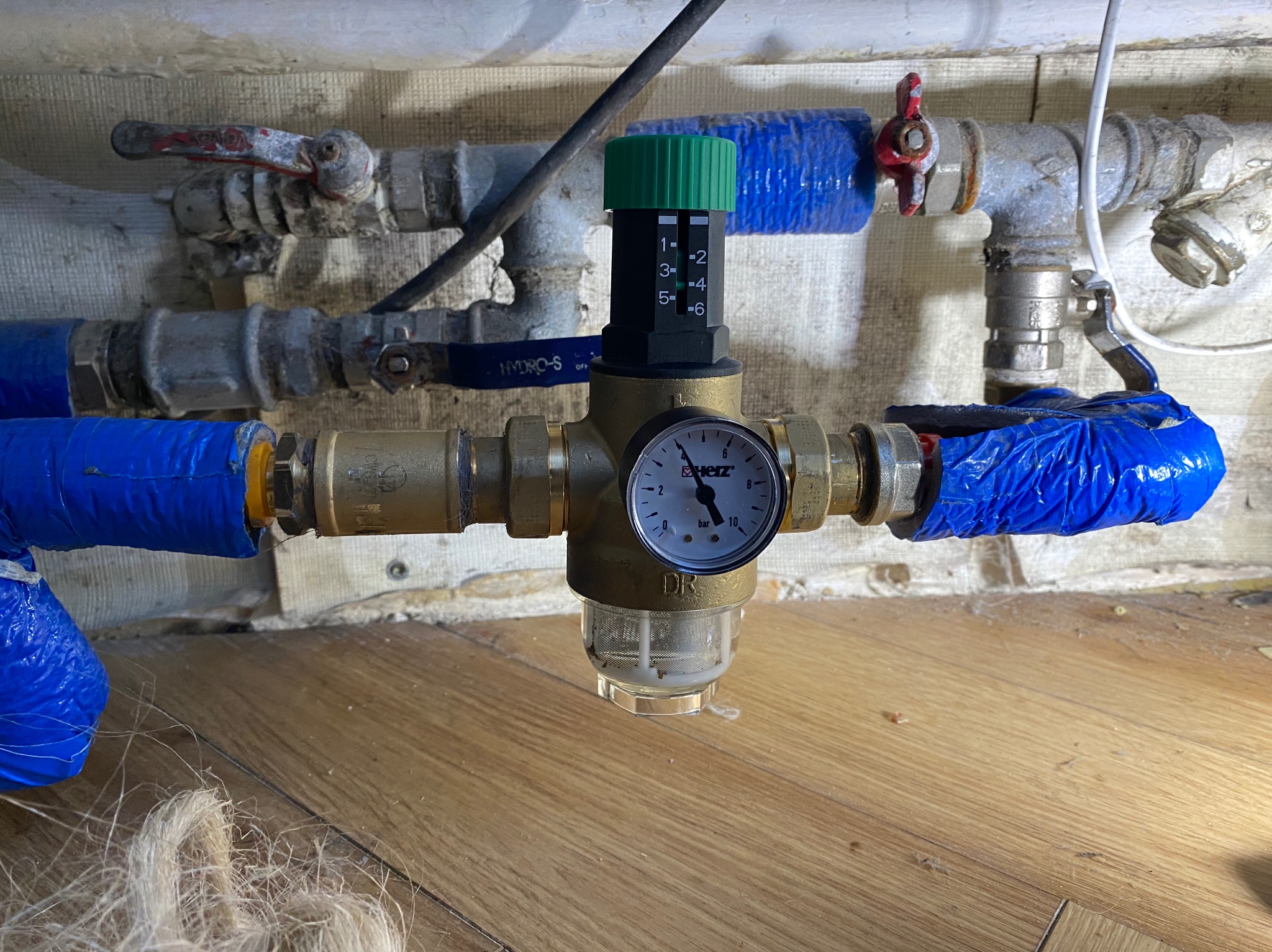 Zamontowany reduktor ciśnienia wody z manometrem i filtrem, widoczne rury z zaworami. Instalacja hydrauliczna w pomieszczeniu z drewnianą podłogą i wykończoną ścianą.