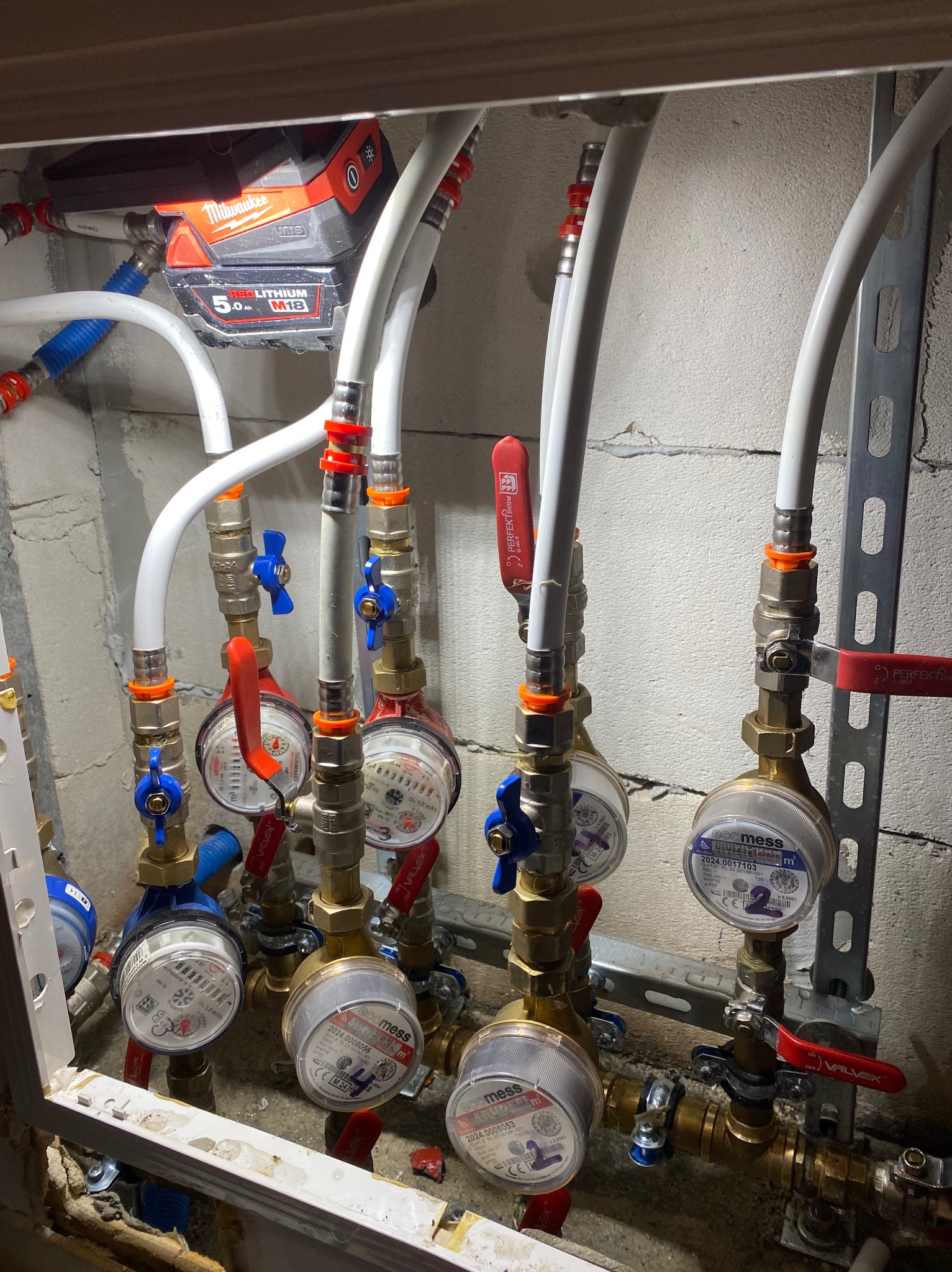 Węzeł wodomierzowy z licznymi zaworami i rurami, zamontowany w przestrzeni pod zabudową. Widoczne liczniki wody, białe rury oraz bateria Milwaukee na górze.