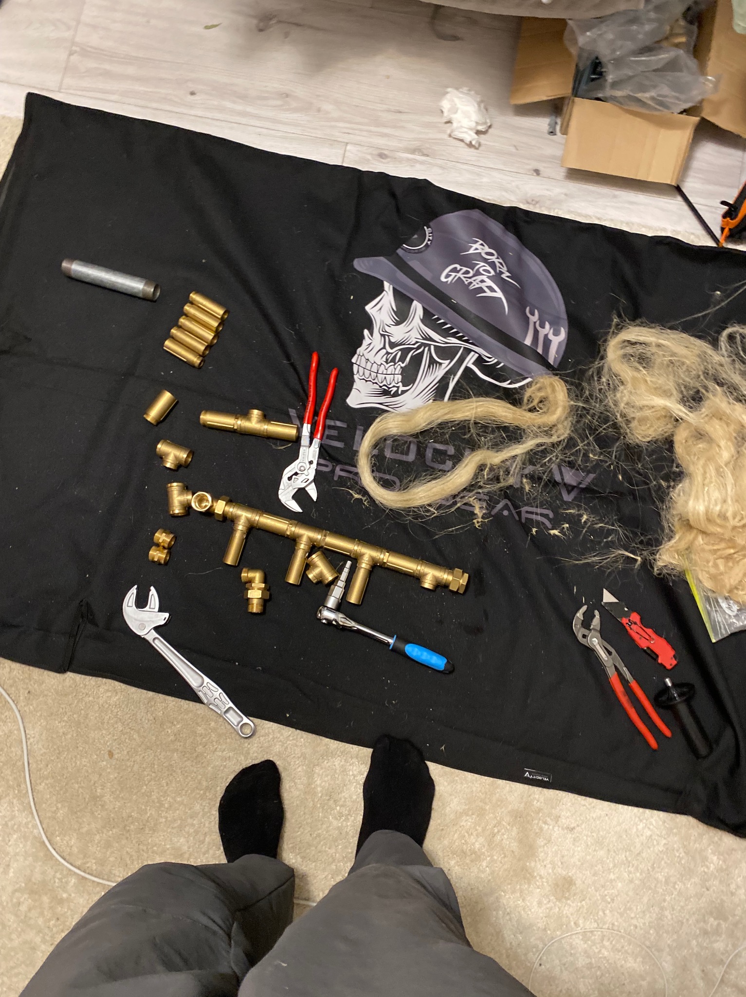 Mosiężne złączki hydrauliczne, klucze i narzędzia na czarnej macie z motywem czaszki. Widoczne stopy w skarpetkach i spodniach. Przygotowanie do montażu instalacji.