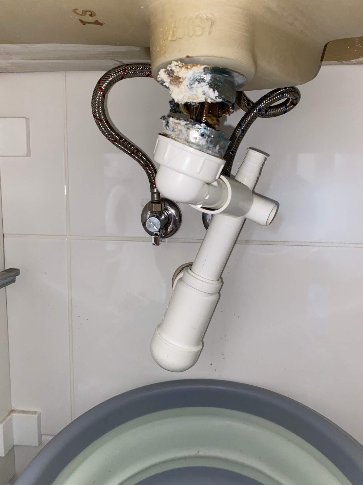 Nieszczelny syfon umywalki z widocznymi śladami korozji i wycieków, pod spodem wiadro. Ujęcie z dołu, na białym tle płytek.