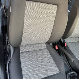 Tamara - Brudne, szare siedzenie samochodowe z plamami przed czyszczeniem, widoczny fragment pasów bezpieczeństwa i dźwigni hamulca ręcznego.