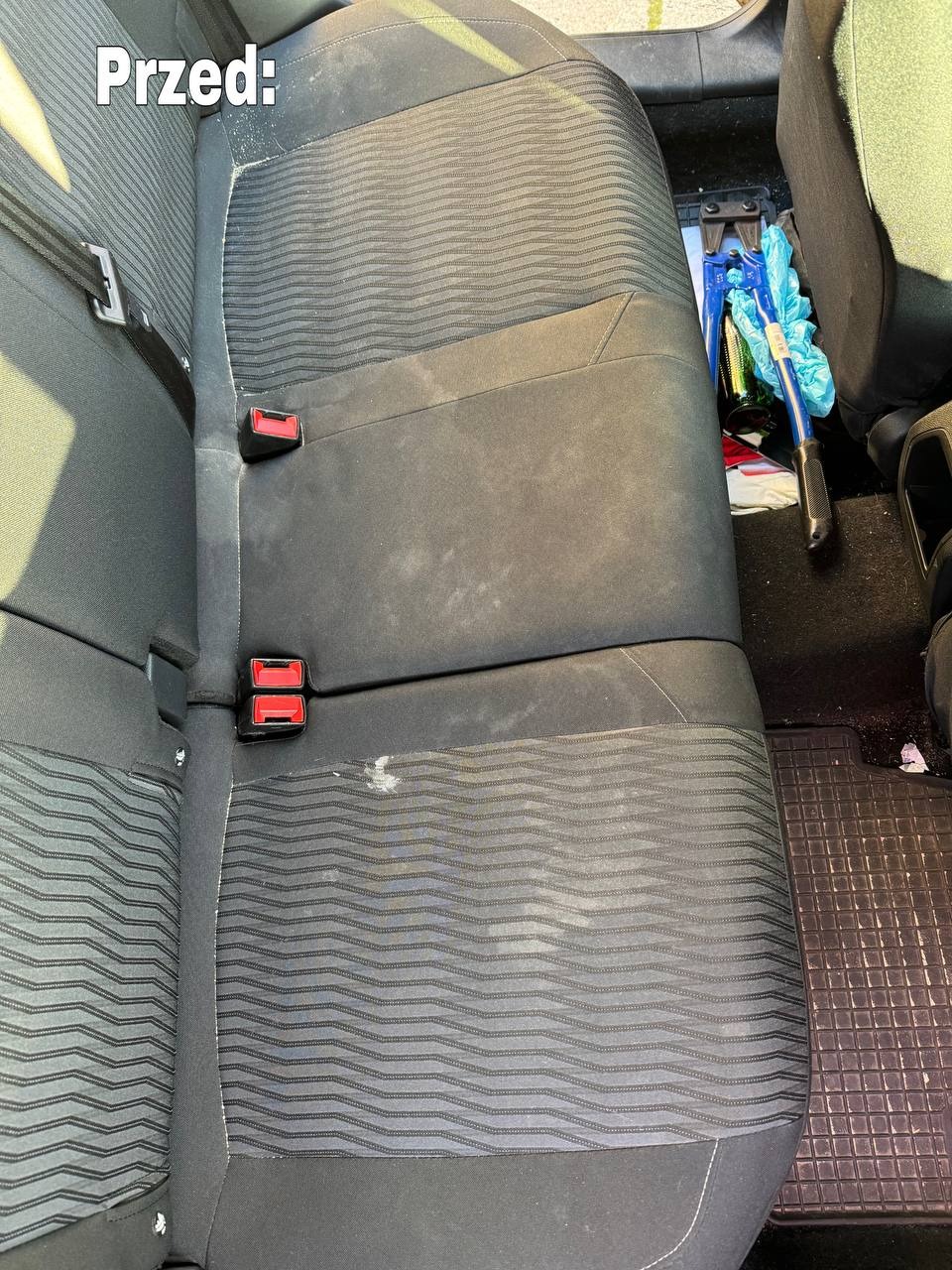Zabrudzona, tylna kanapa samochodowa z widocznymi plamami i napisem 'Przed', obok leżą narzędzia i akcesoria do czyszczenia.